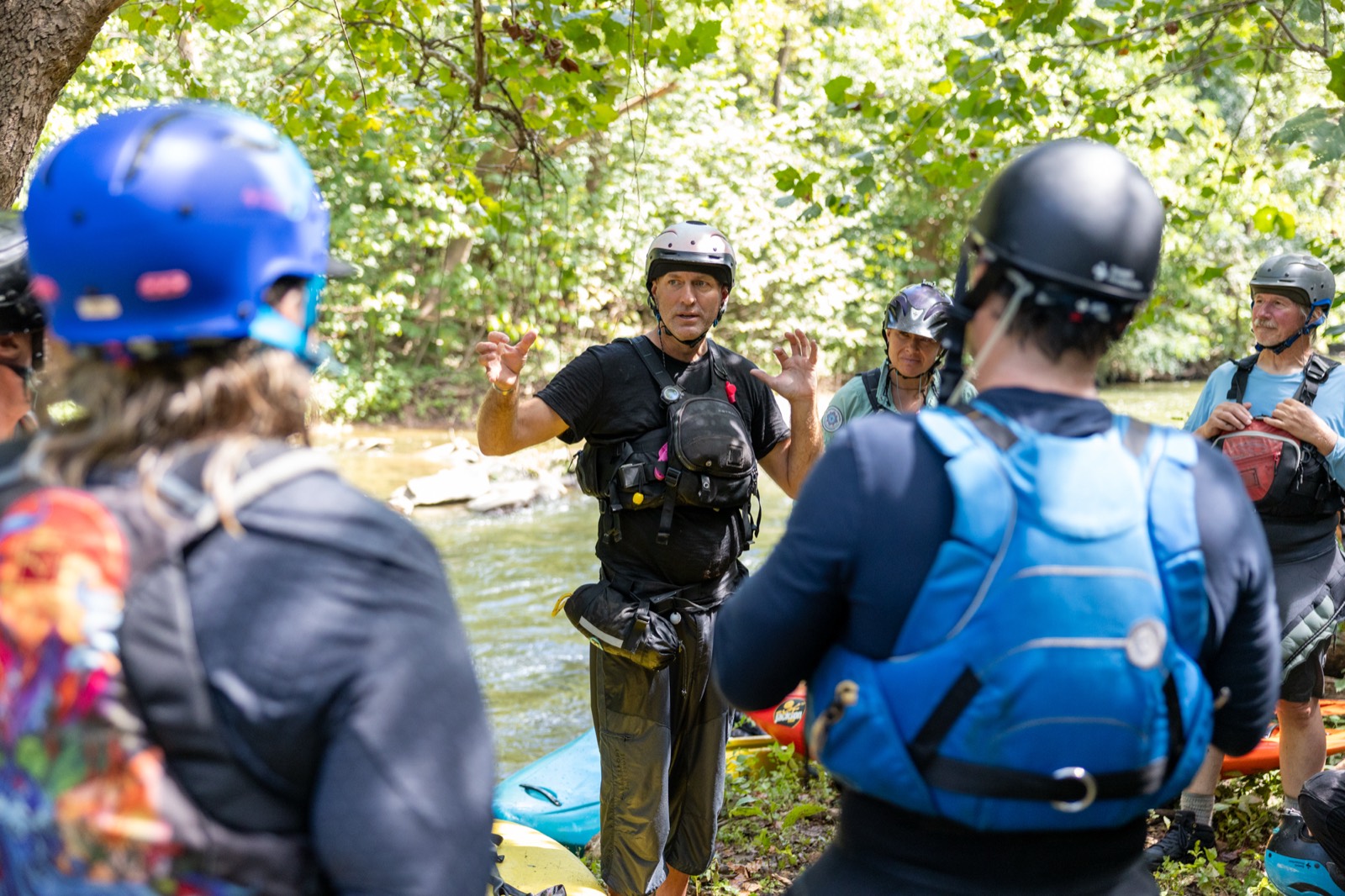 An instructor in a helmet and rescue vest talks with a group of paddlers on a wooded riverbank.